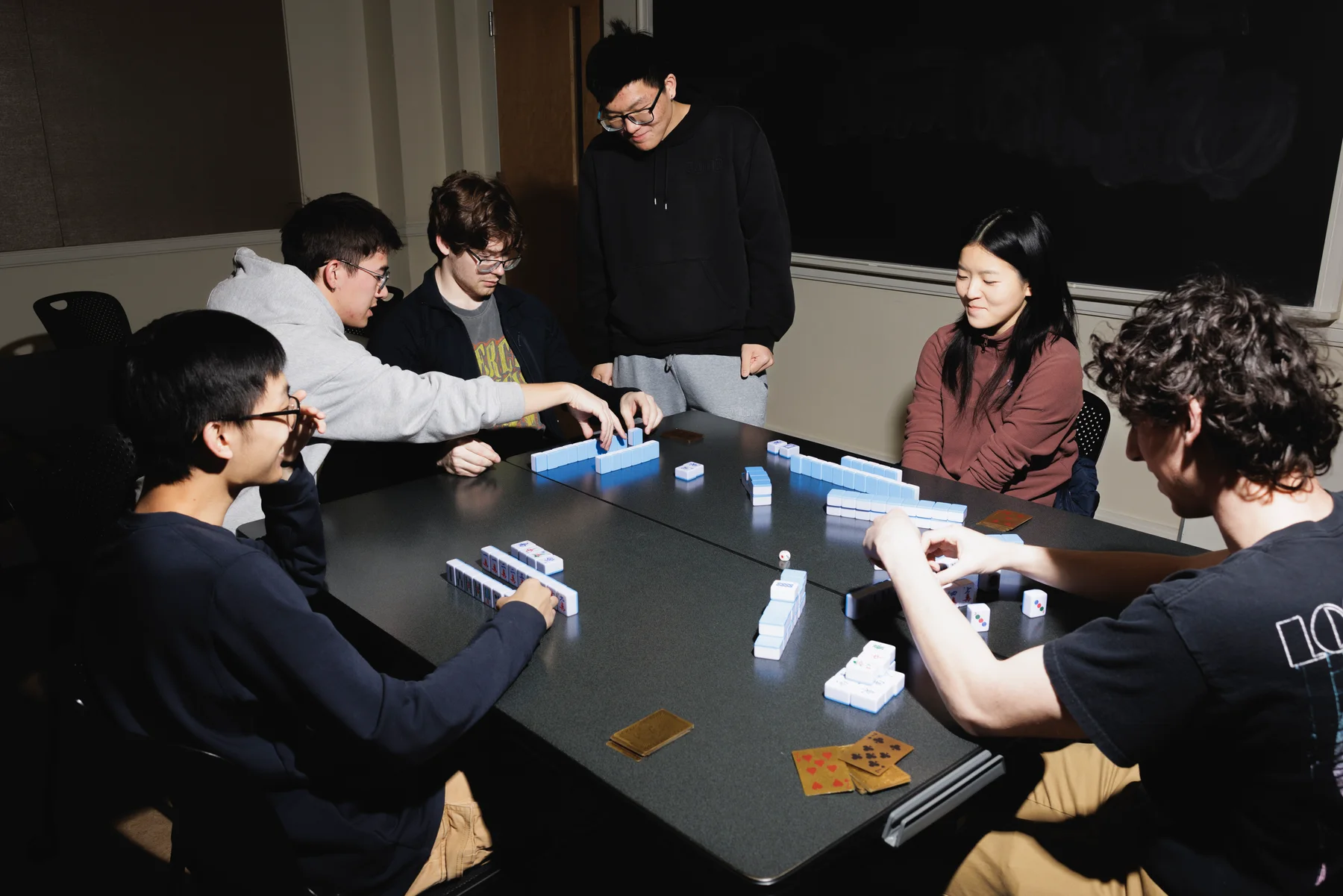 Club members gathered around a table playing mahjong with blue-backed tiles.